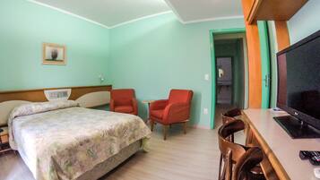 Quarto de hotel com uma cama, duas cadeiras vermelhas, uma mesa de trabalho com uma televisão e uma porta que dá para outro quarto.