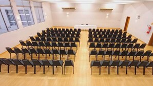 Un salón de conferencias con filas de sillas negras, una mesa blanca al frente y ventanas amplias a un lado.