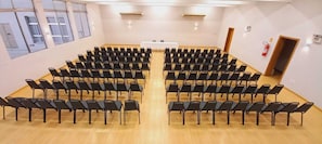 Un salón de conferencias con filas de sillas negras, una mesa blanca al frente y ventanas amplias a un lado.