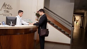 Una recepción de hotel con un mostrador de madera y un monitor de computadora. Se observa una señal con el nombre del hotel sobre el mostrador. Una escalera es visible al fondo.