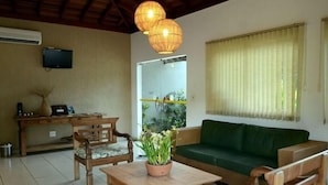 A room with a green sofa, wooden chairs, a table with a potted plant, a TV on the wall, and a window with curtains. - Pousada Vitalis (Olímpia)