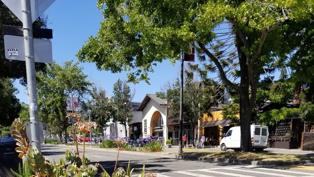 Una calle con un paso de peatones, una camioneta blanca estacionada al costado y un edificio con una entrada arqueada al fondo.
