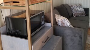 Un espacio habitable compacto con área de kitchenette que incluye un microondas, una cafetera y una cesta de mimbre sobre una repisa de vidrio. Un sofá gris con cojines estampados está ubicado contra la pared.