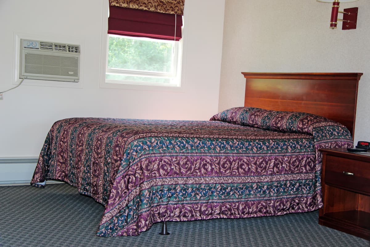 Une chambre à coucher comprenant un lit, une table de chevet, une fenêtre agrémentée d’un lambrequin rouge, un appareil de climatisation et un luminaire fixé au mur.