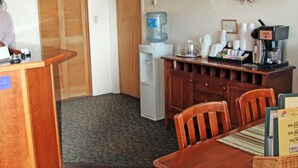 A hotel room with a wooden desk, a small kitchenette, a dining area with a table and chairs, and a water dispenser.