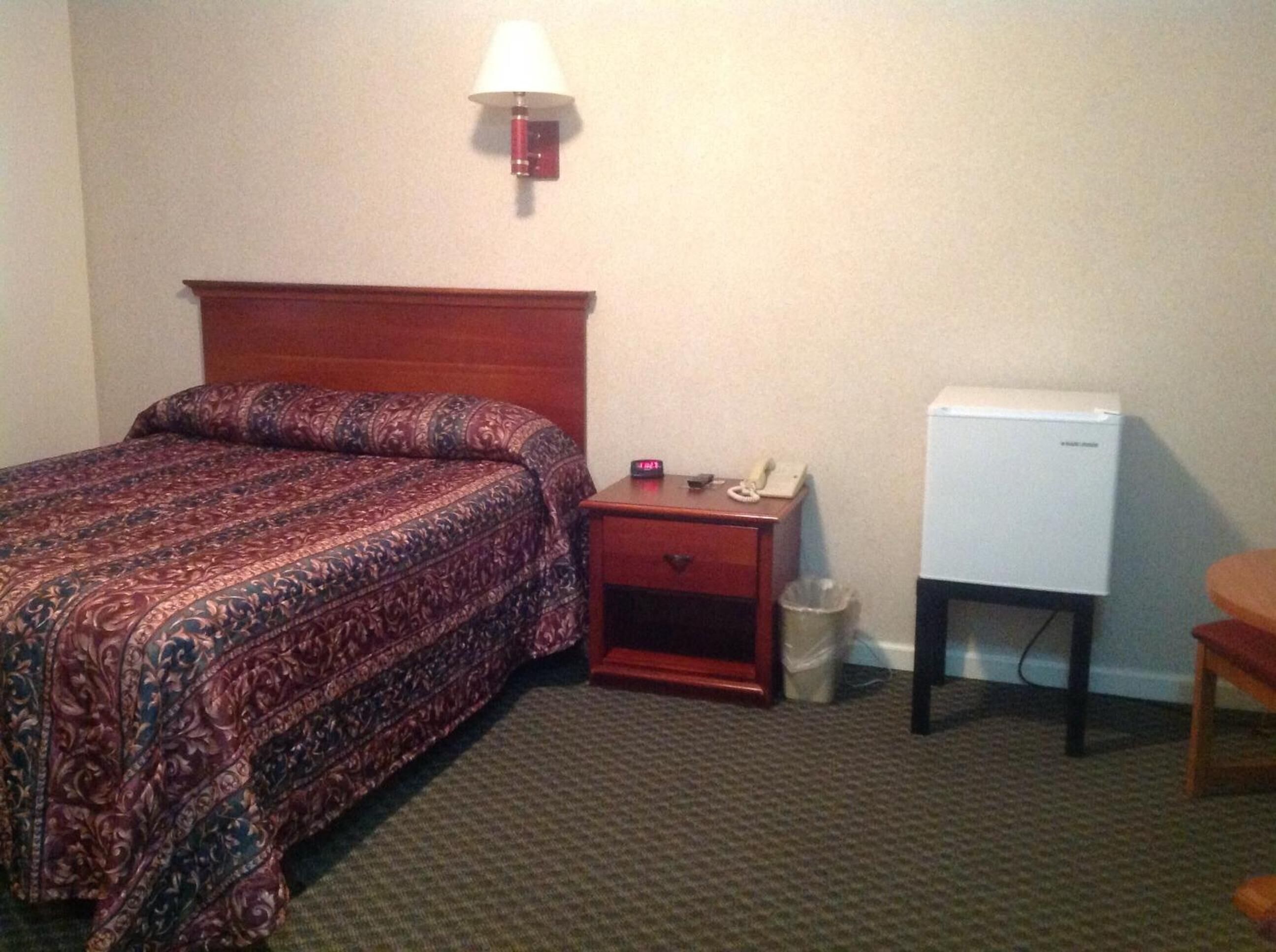 A hotel room with a bed, a nightstand with a lamp, a telephone, and a small refrigerator.