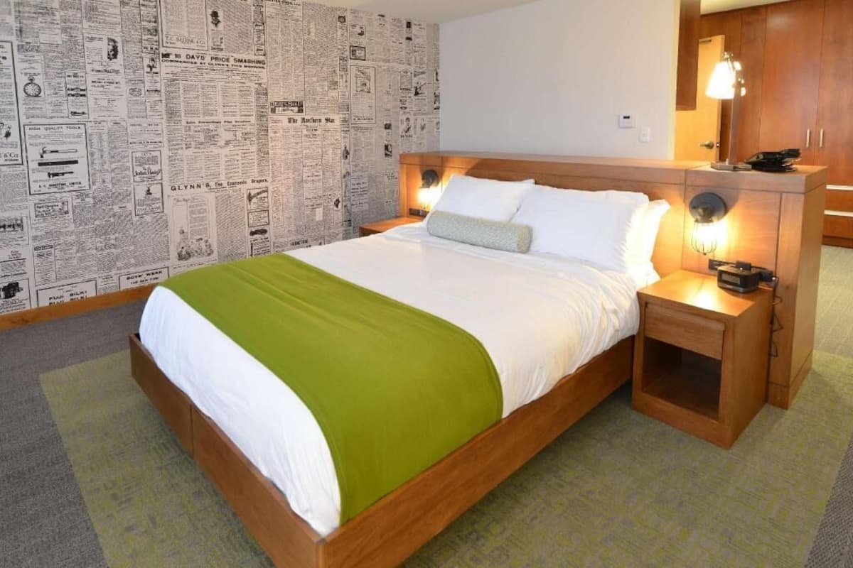 A hotel room with a bed, wooden headboard, bedside tables, a lamp, and a wall covered with newspaper clippings.