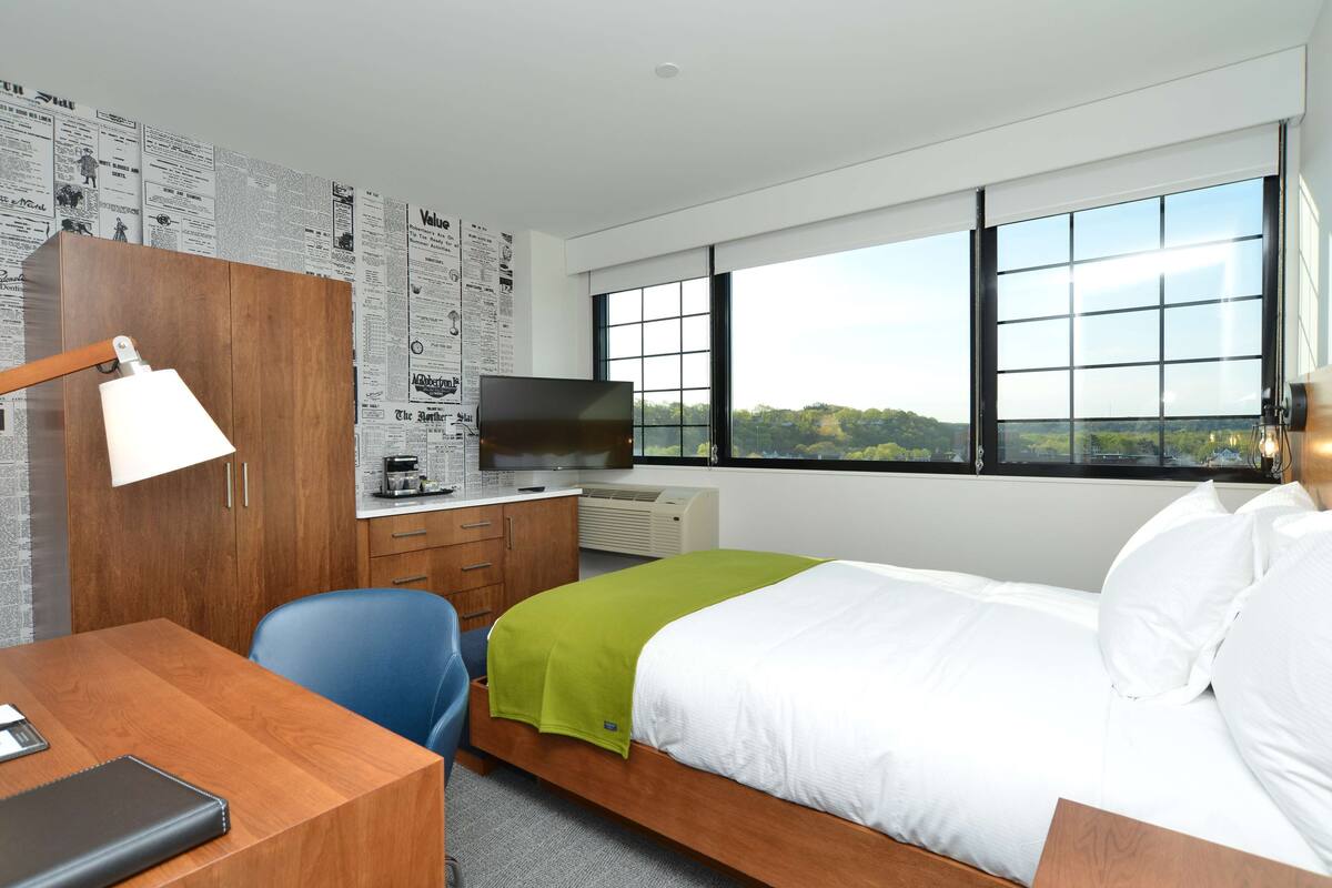 A modern hotel room with a wooden desk, a television, a bed with white and green bedding, and large windows offering a view of the outdoors.
