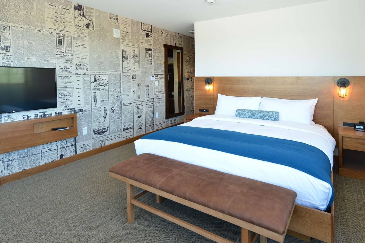 A hotel room with a large bed, a bench, a TV, and a wall covered with newspaper clippings.
