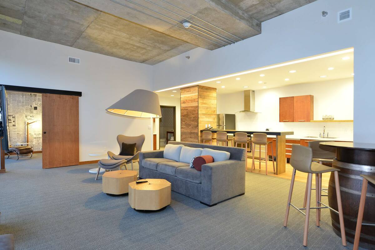 A modern living room with a grey sofa, round ottoman, and a lamp. A kitchen area with wooden cabinets and a bar counter is visible in the background.
