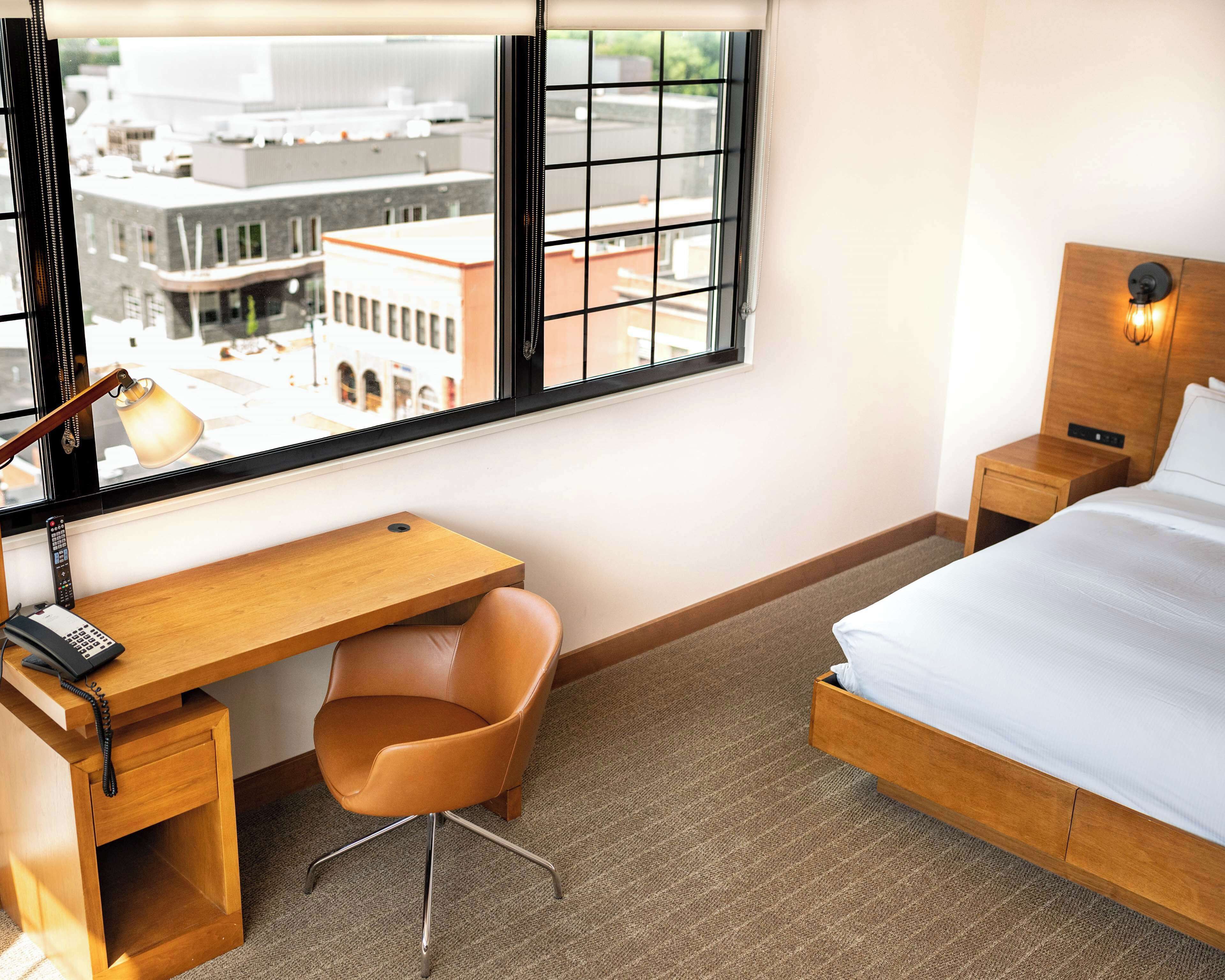 A hotel room with a wooden desk, a chair, a bed, a nightstand, a lamp, and a view of the city through a window.