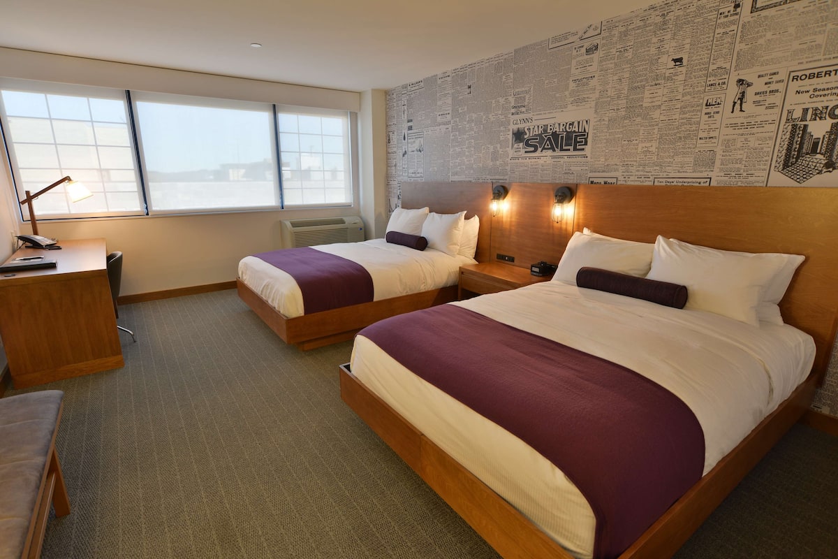 A hotel room with two beds, a desk, and a large window.