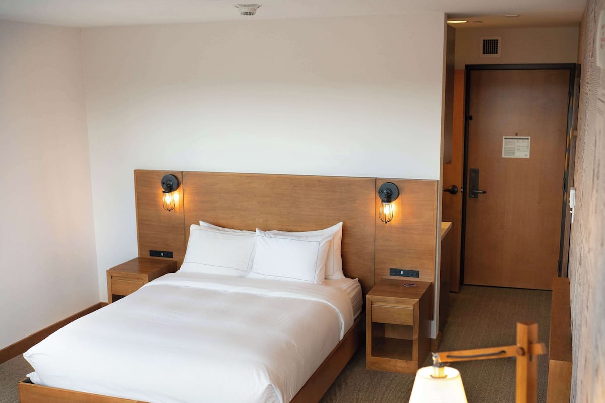 A hotel room with a wooden bed, two bedside tables with lamps, a wooden door, and a carpeted floor.