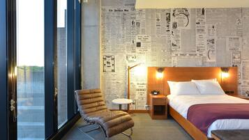 Ein Hotelzimmer mit großem Fenster, einem Bett mit Holz-Kopfteil, einem Ledersessel, einem kleinen runden Tisch und einem Nachttisch mit Lampe.