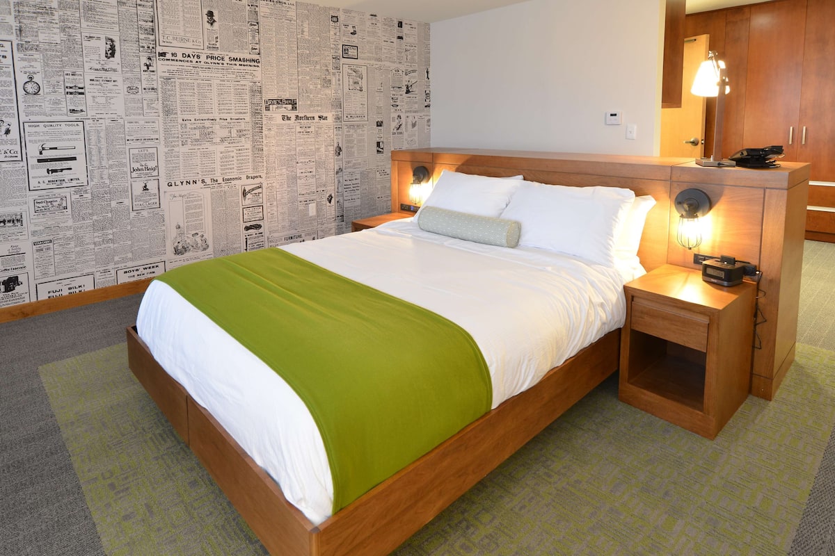 A hotel room with a wooden bed, green and white bedding, a nightstand with a lamp, and a wall covered with newspaper clippings.