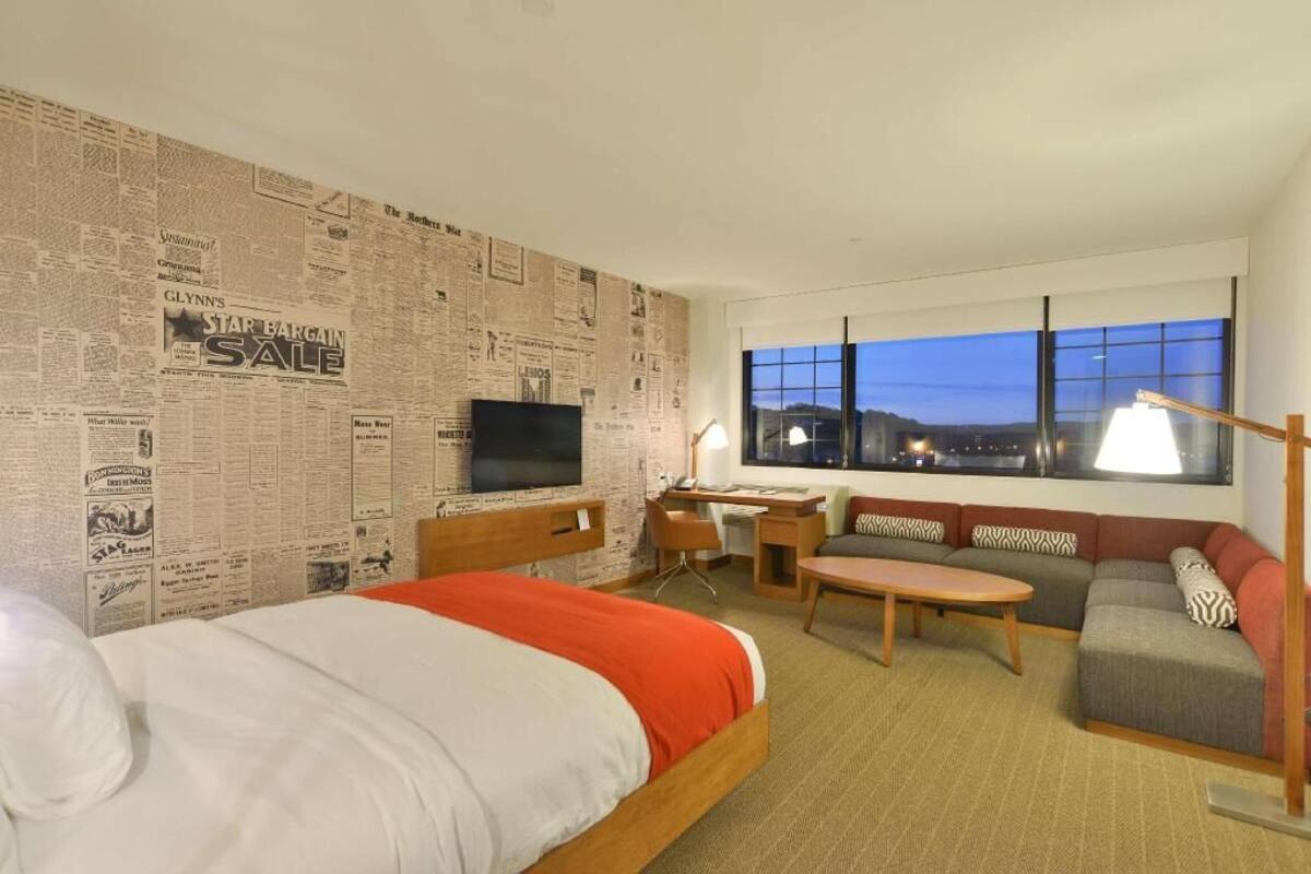 A hotel room with a large bed, a desk, a TV, a sofa, and a window with a city view.