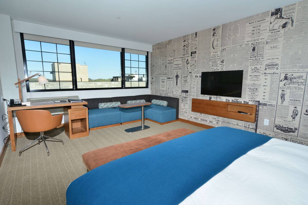 A modern hotel room with a large bed, a desk with a chair, a sofa, a television, and a large window offering a city view.