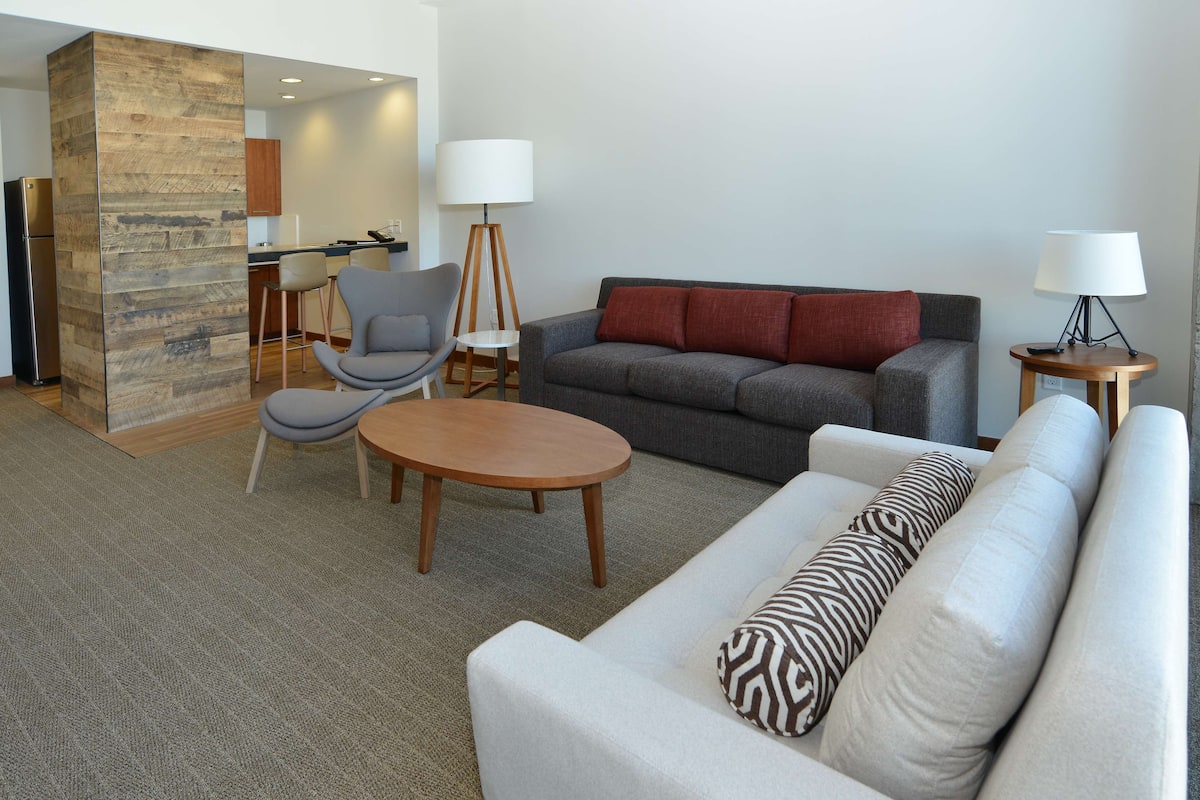 A modern living room with a grey sofa, a white sofa, a wooden coffee table, a grey armchair, and a small round table with a lamp.
