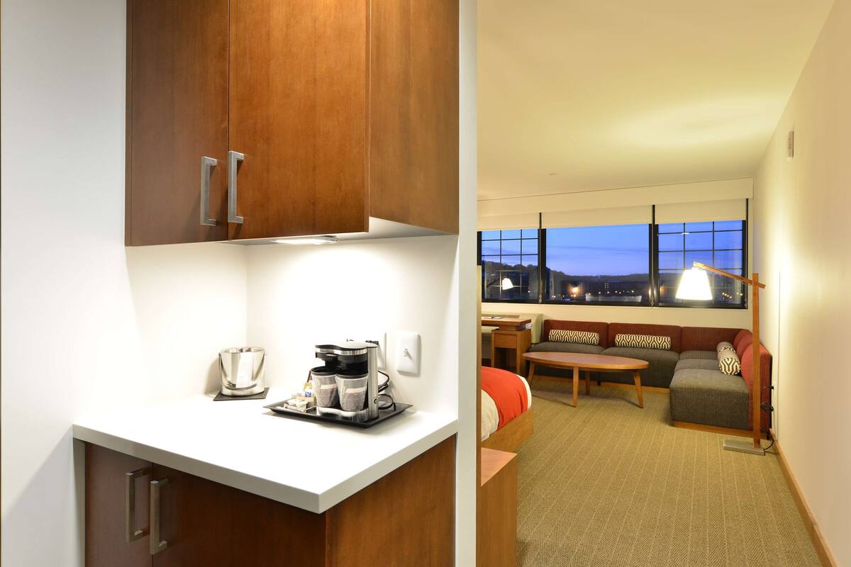 A hotel room with a kitchenette, a seating area with a sofa and armchairs, and a large window offering a city view.