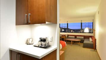 A hotel room with a kitchenette, a seating area with a sofa and armchairs, and a large window offering a city view.