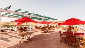 A covered outdoor seating area with red umbrellas and wooden chairs, overlooking a scenic landscape. - Hôtel Taghit Saoura (Taghit)