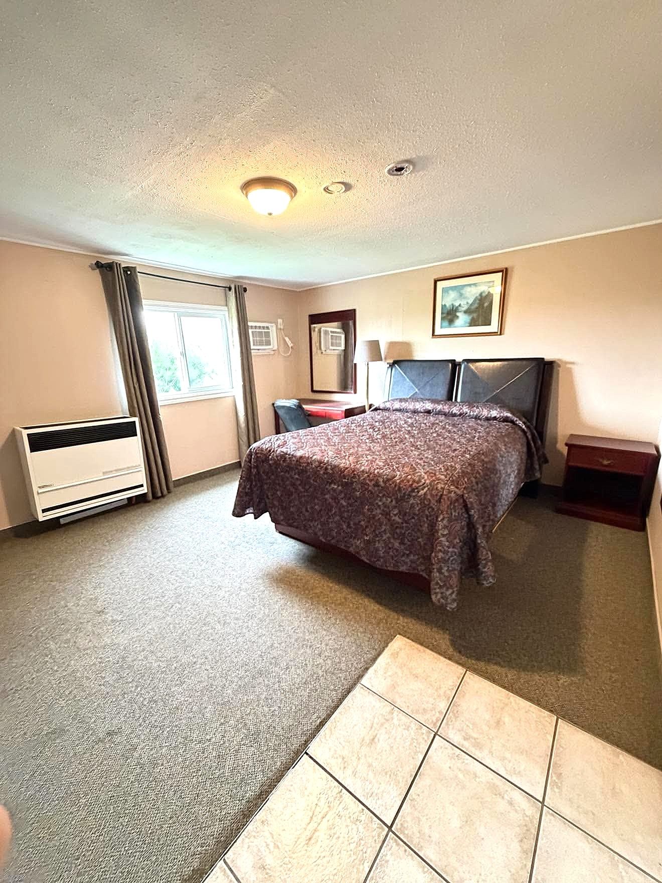 Habitación de hotel con cama, escritorio, silla, calefactor, ventana con cortinas y un cuadro en la pared.