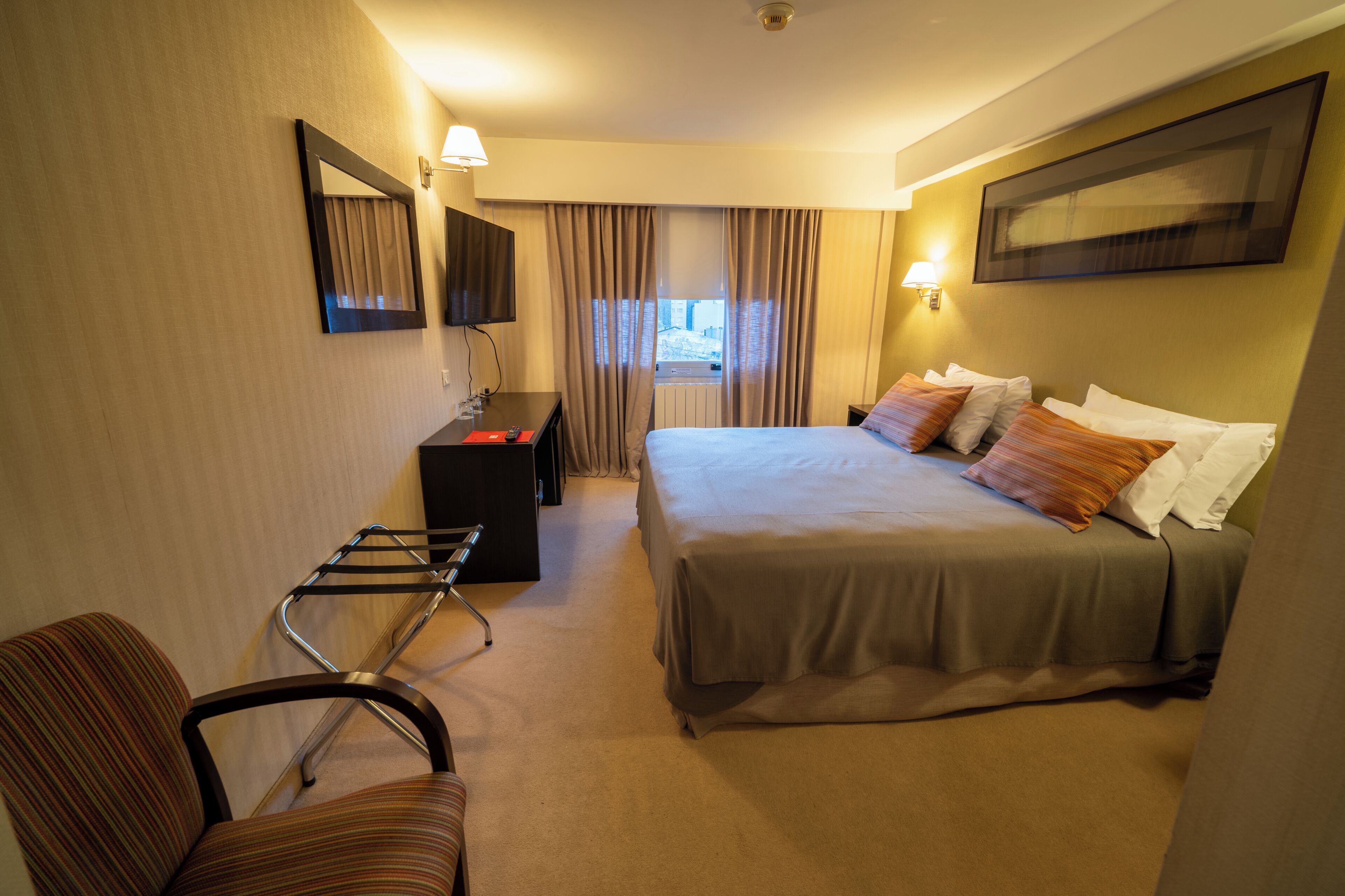 Habitación de hotel con cama, escritorio, silla, ventana con cortinas y televisor montado en la pared.