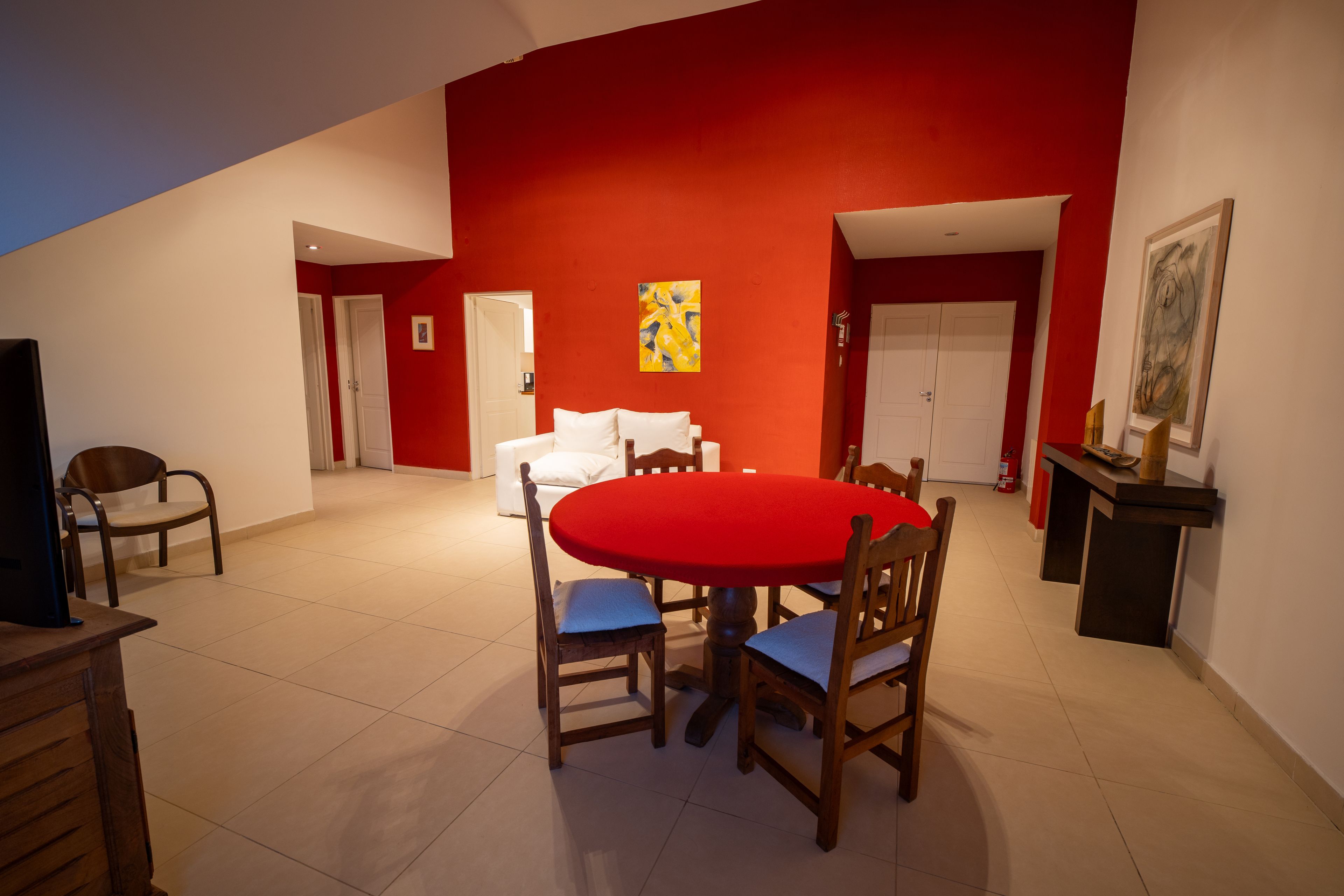 A dining area with a red table and chairs, white walls, and a painting on the wall.