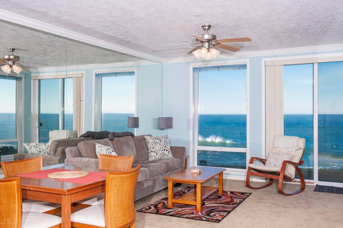 A spacious living room with a large view of the ocean, a ceiling fan, and a seating area with a table and chairs.