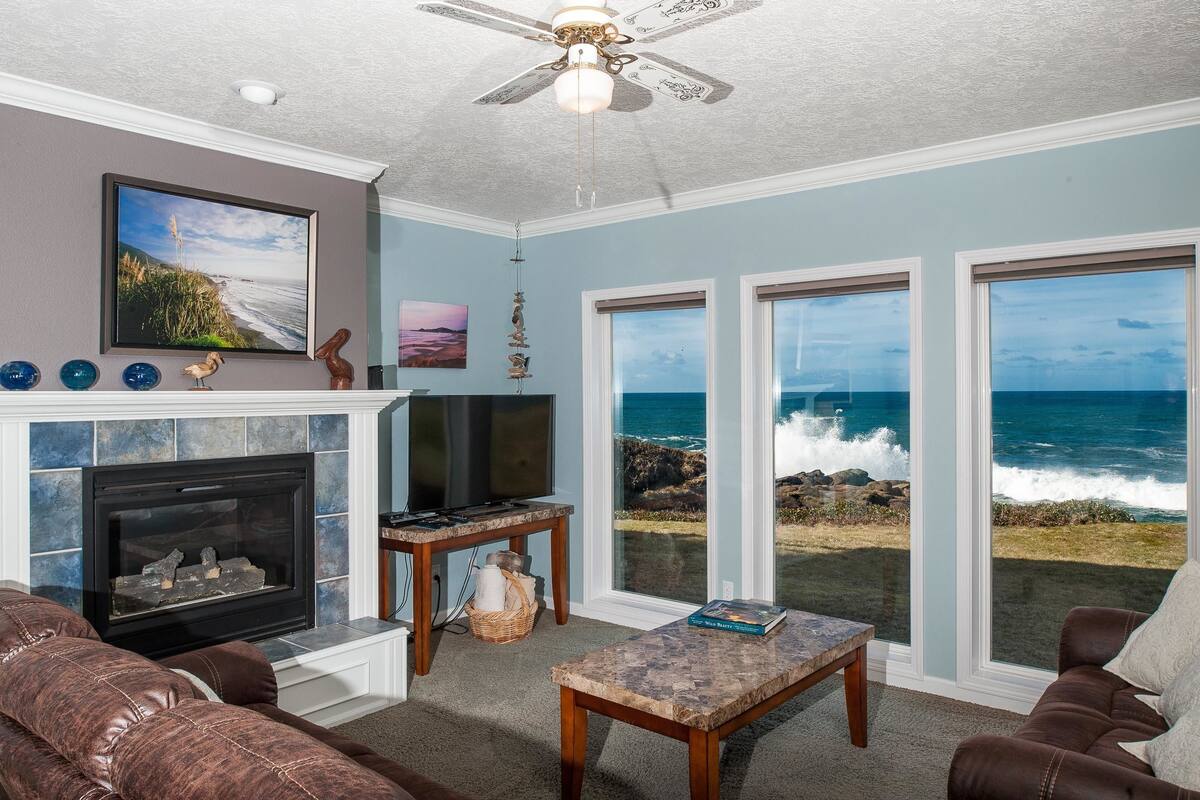 A living room with a fireplace, a television, a ceiling fan, and large windows overlooking the ocean.