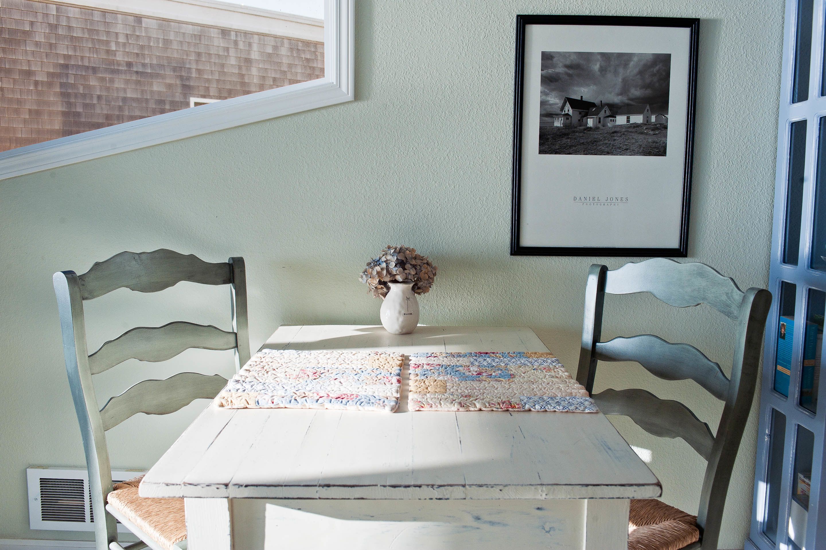 Una mesa con un jarrón de flores secas, dos sillas y una fotografía enmarcada en la pared.