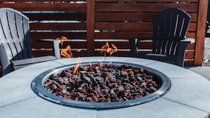Un hoyo para fuego al aire libre de forma redonda, con carbón encendido, rodeado por una mesa de piedra y dos sillas Adirondack.