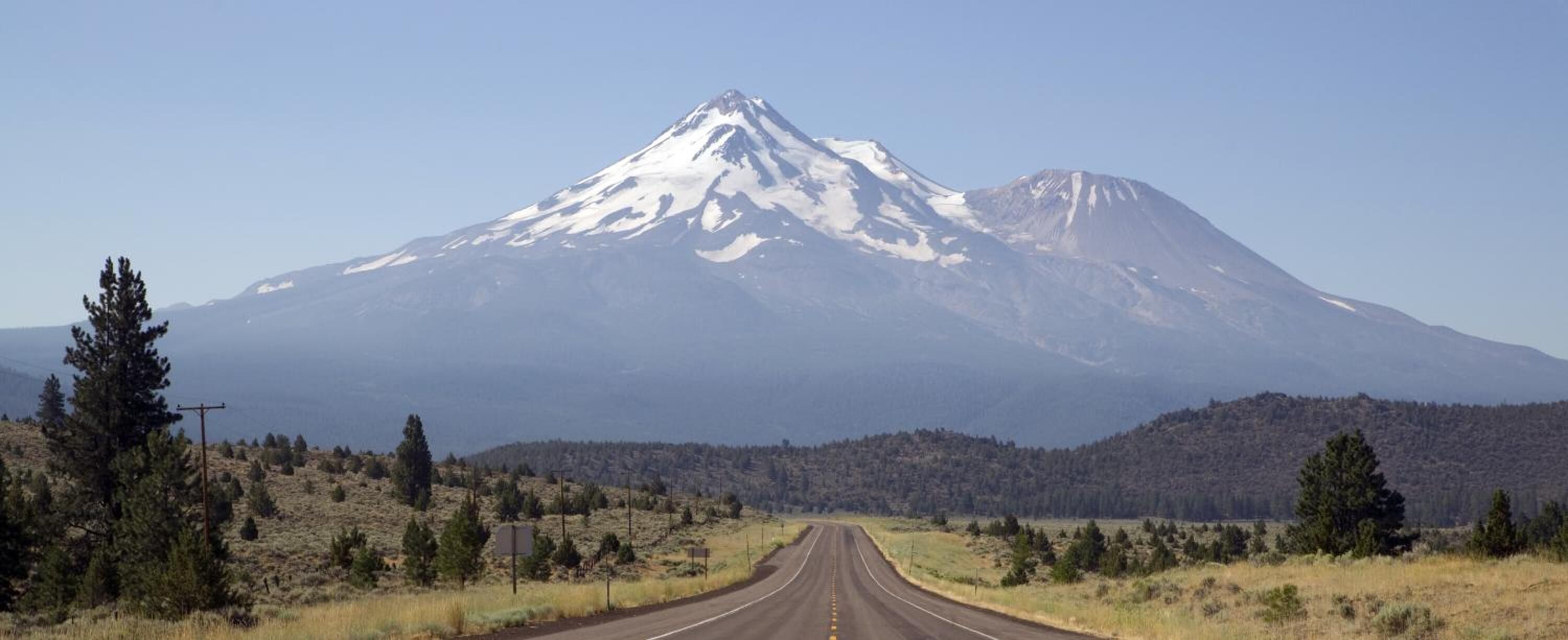 A road leading to a snow-capped mountain with a forested hill in the background.