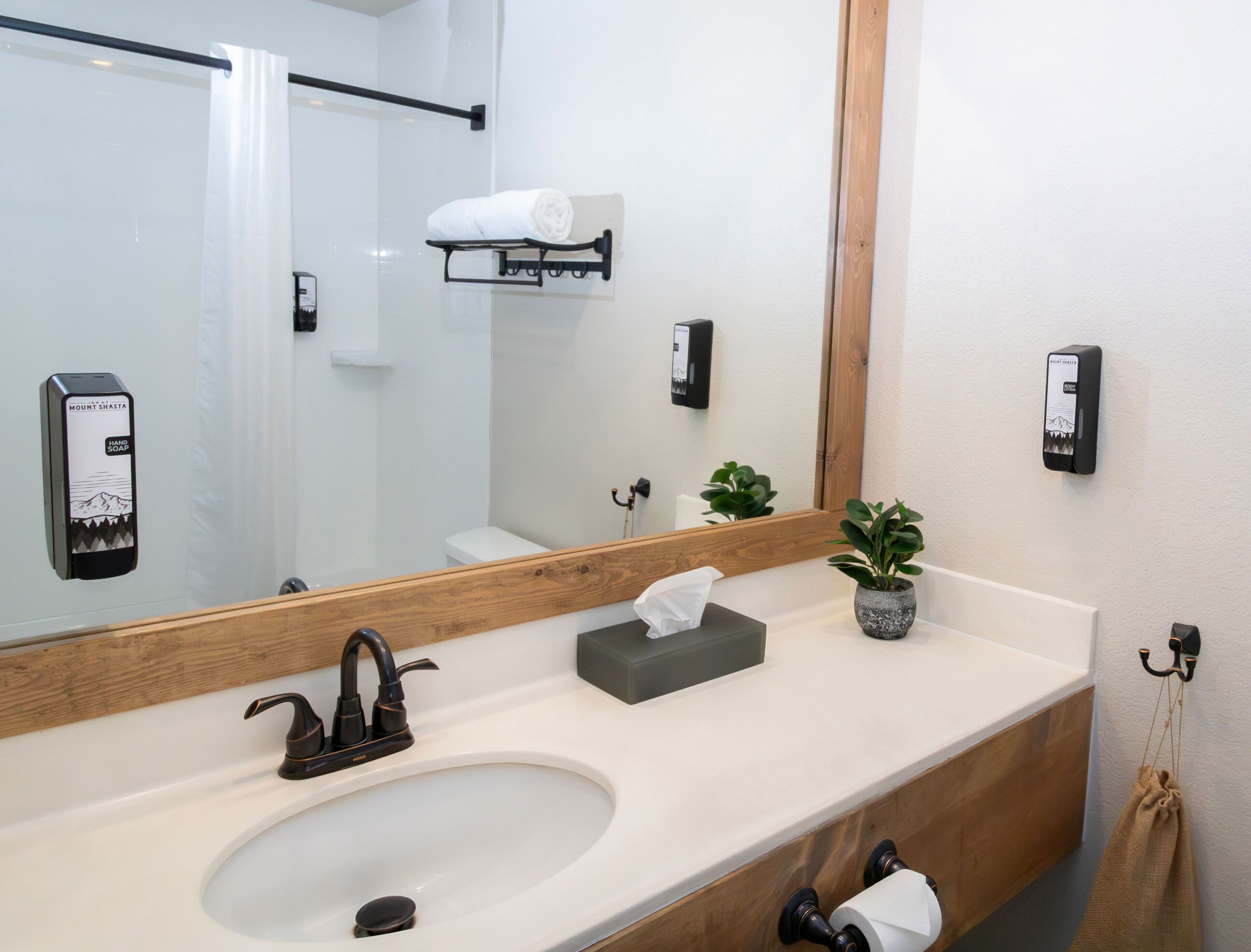 A modern bathroom with a large mirror, a sink, a towel rack, a soap dispenser, and a small potted plant.