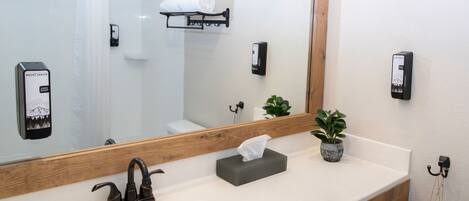 A modern bathroom with a large mirror, a sink, a towel rack, a soap dispenser, and a small potted plant.