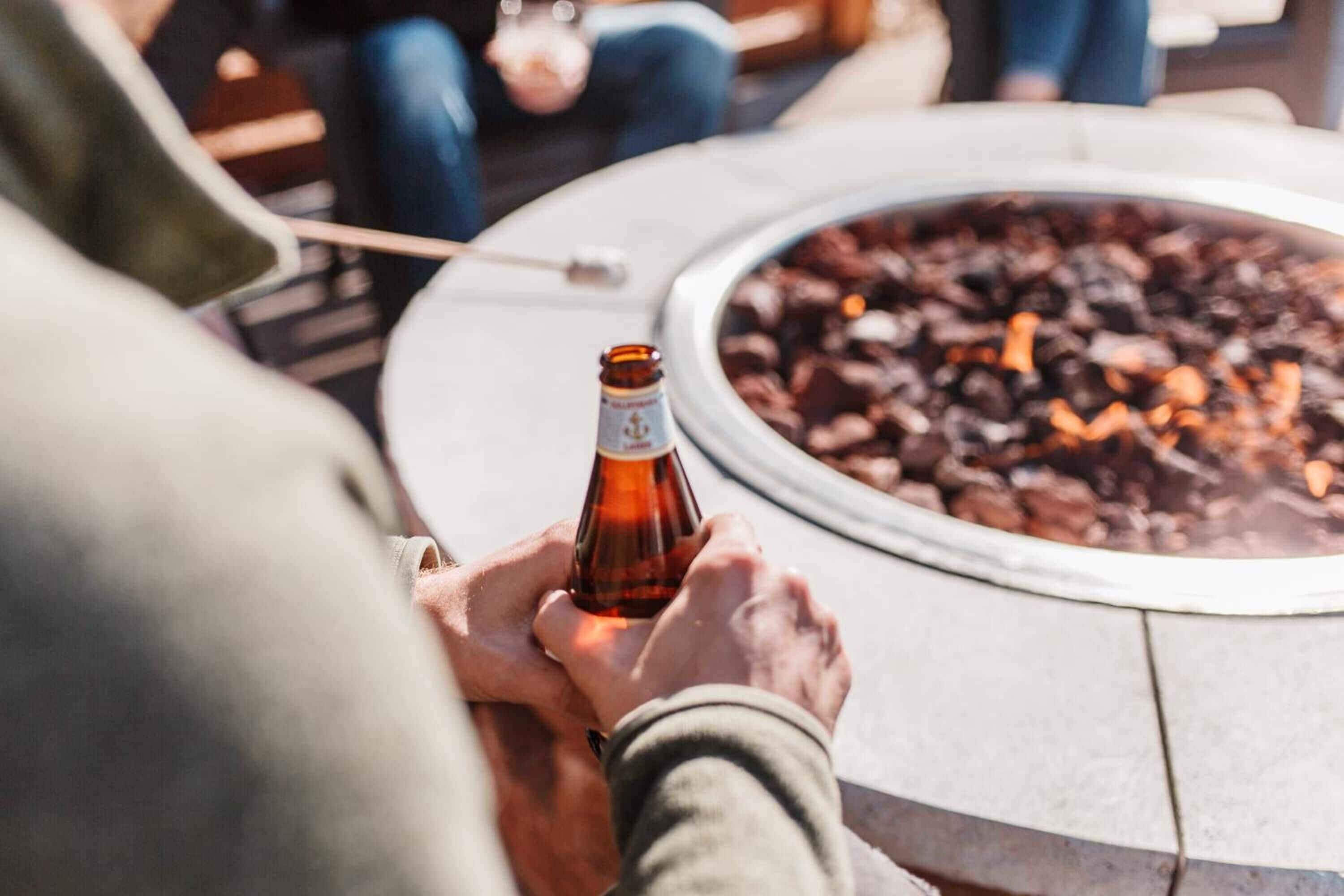 A person holding a beer bottle at a round outdoor table with a fire pit in the background.