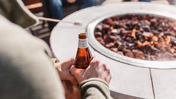 A person holding a beer bottle at a round outdoor table with a fire pit in the background.