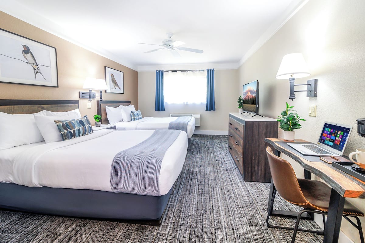 A hotel room with two beds, a desk with a laptop, a chair, a desk lamp, a ceiling fan, and a TV.