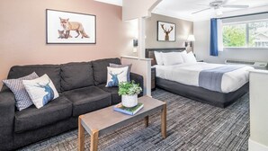 Una habitación de hotel con un sofá gris oscuro, una cama con ropa blanca, un ventilador de techo y una ventana con cortinas.