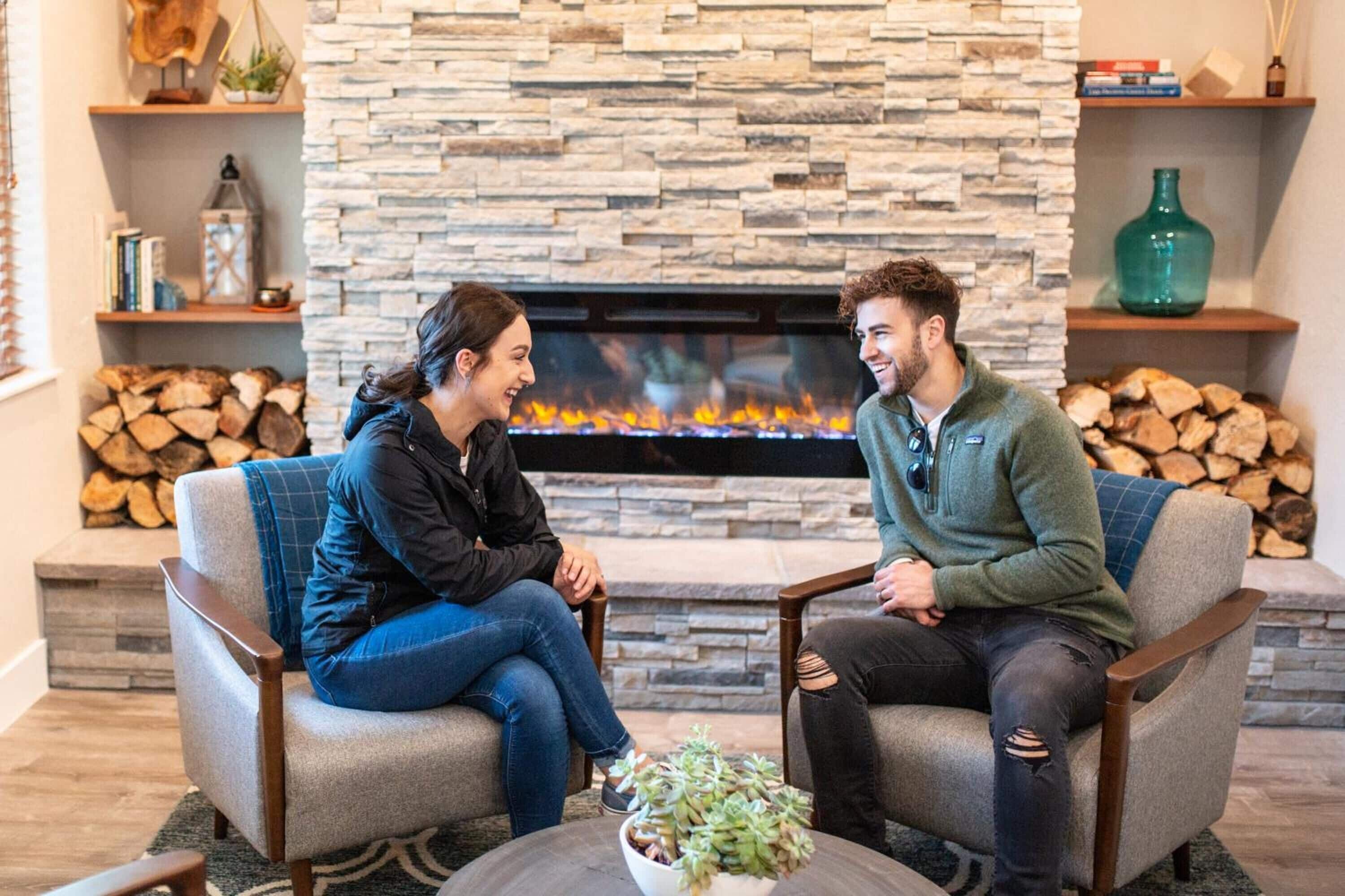 Duas pessoas sentadas em poltronas, uma de frente para a outra, em um ambiente com lareira de pedra, uma mesinha com uma planta e prateleiras com livros e objetos decorativos.