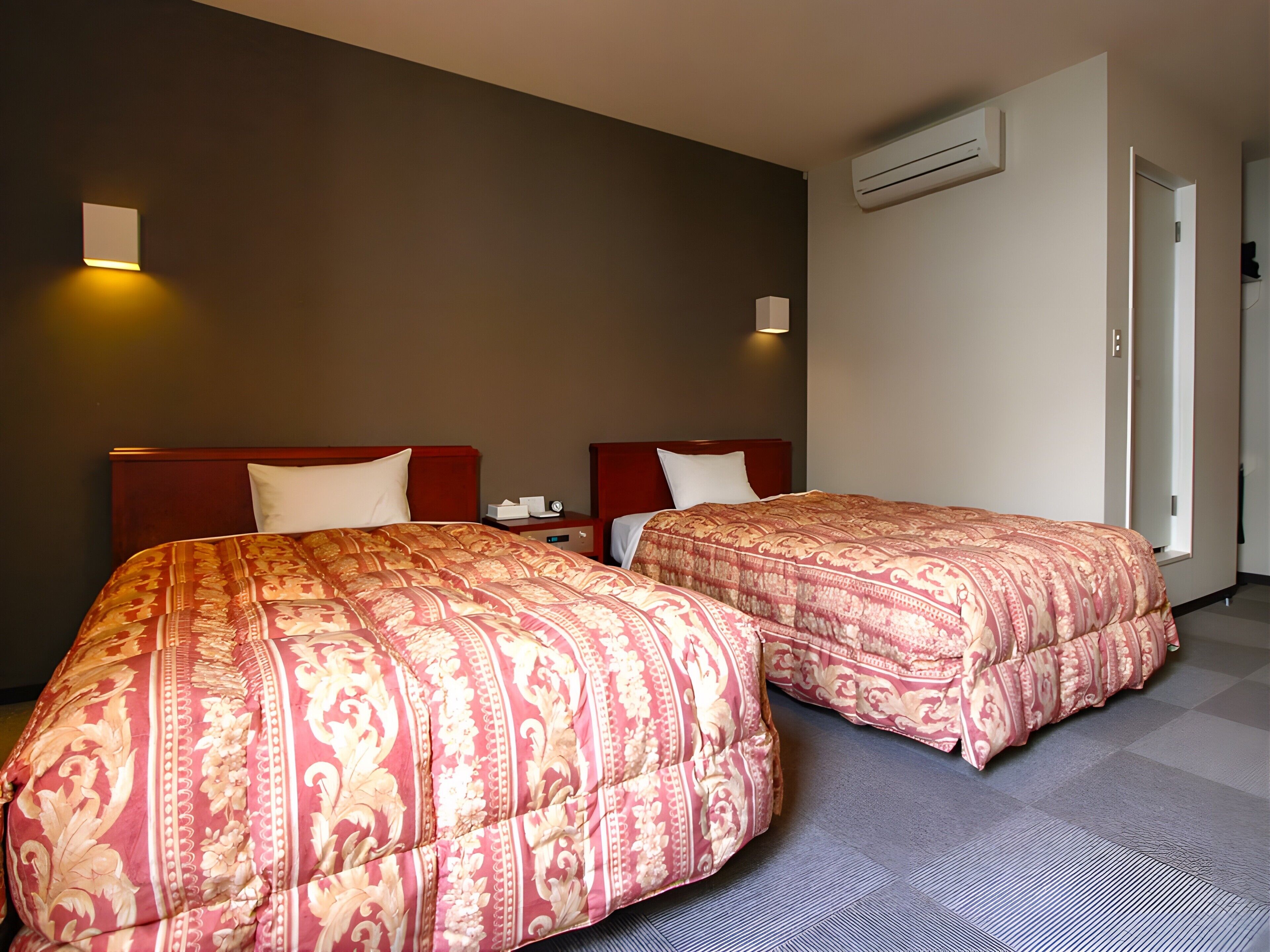 Ein Hotelzimmer mit zwei Betten, jeweils mit einem gemusterten Überwurf, einem Nachttisch mit Lampe und einer Wand-Klimaanlage.