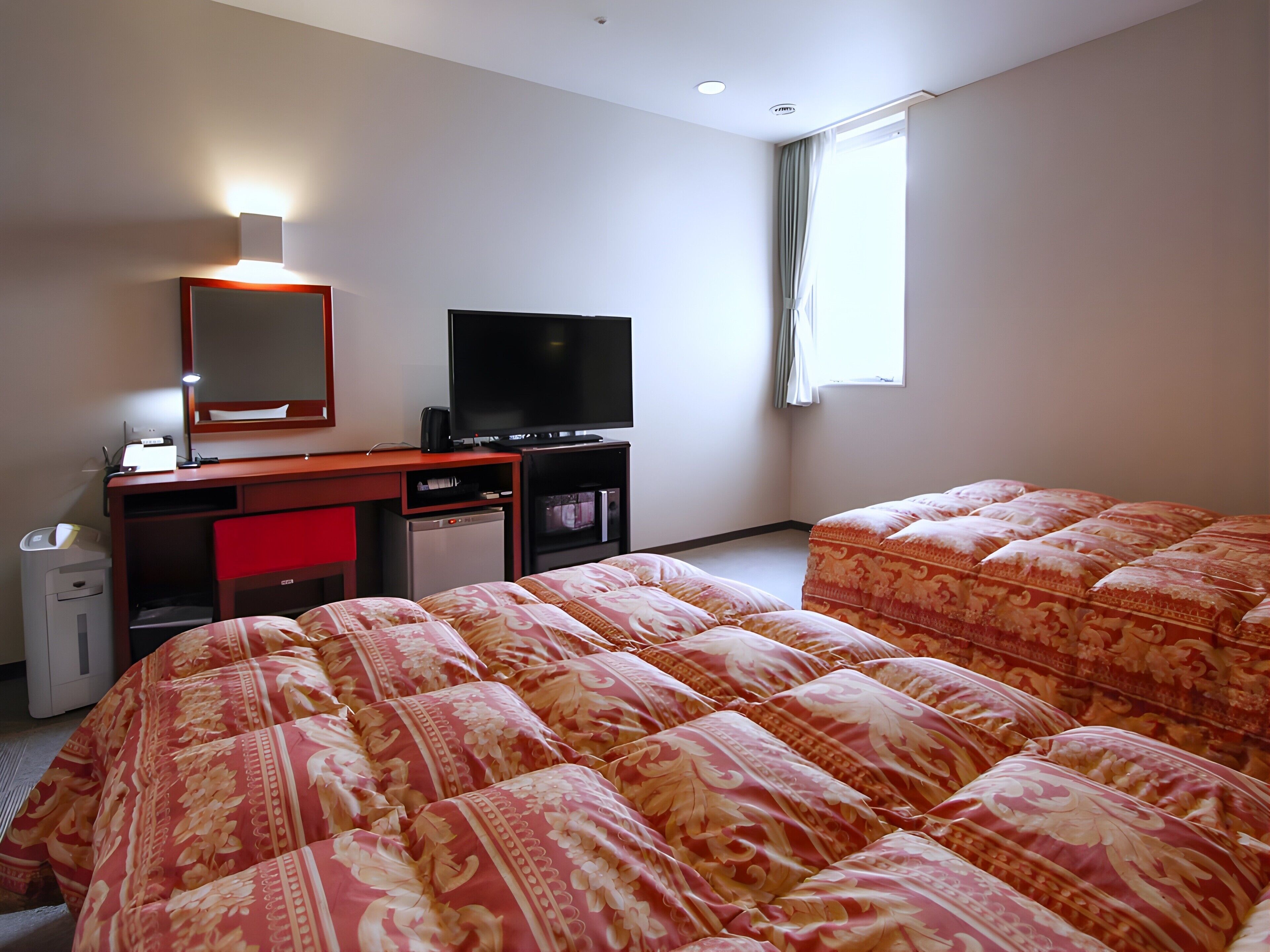 Una habitación de hotel con cama, escritorio con televisor, refrigerador pequeño y ventana con cortinas.