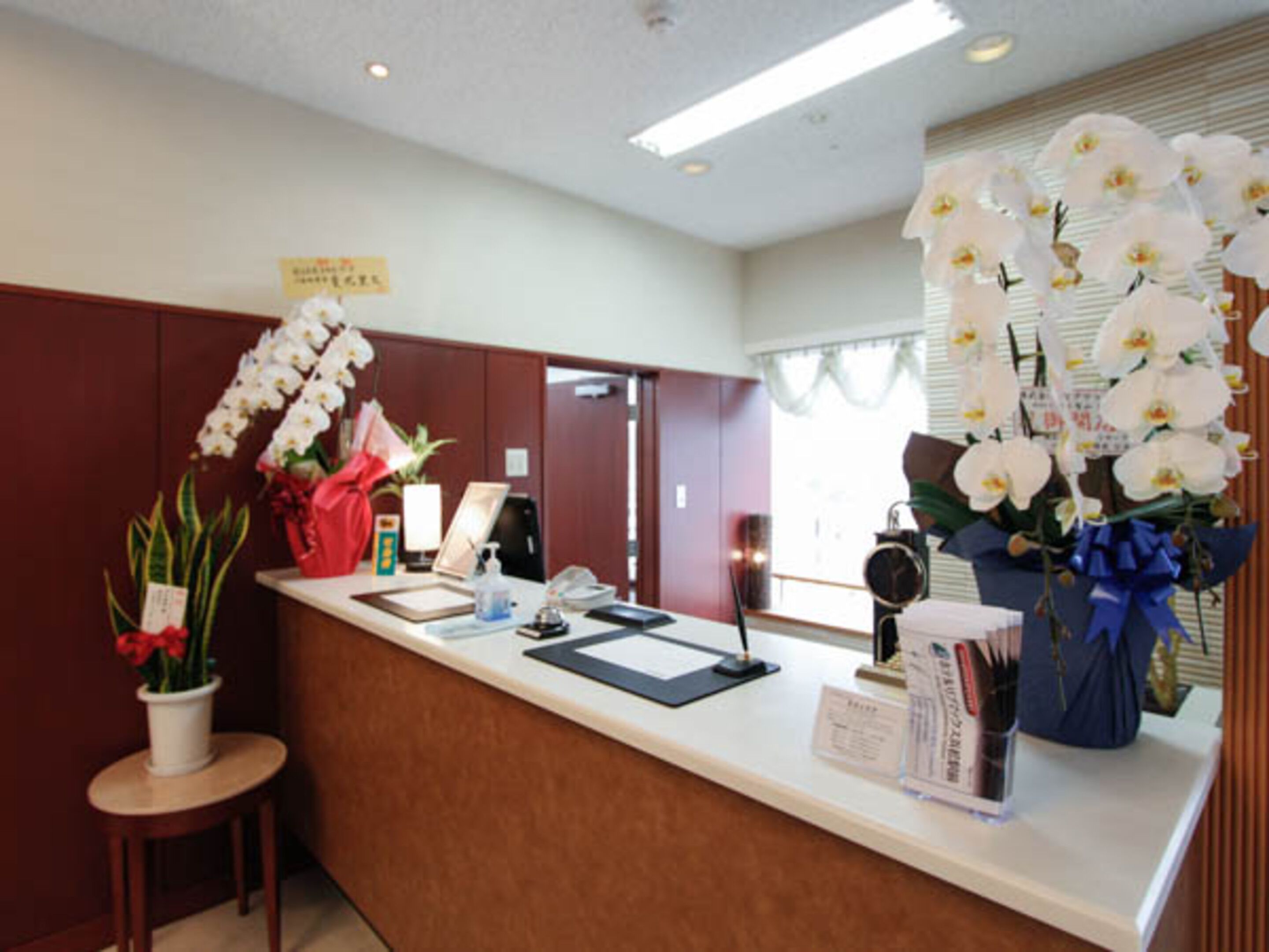 Un mostrador de recepción de hotel con encimera de madera, una pequeña mesa redonda con una planta en maceta y un gran jarrón con orquídeas blancas.