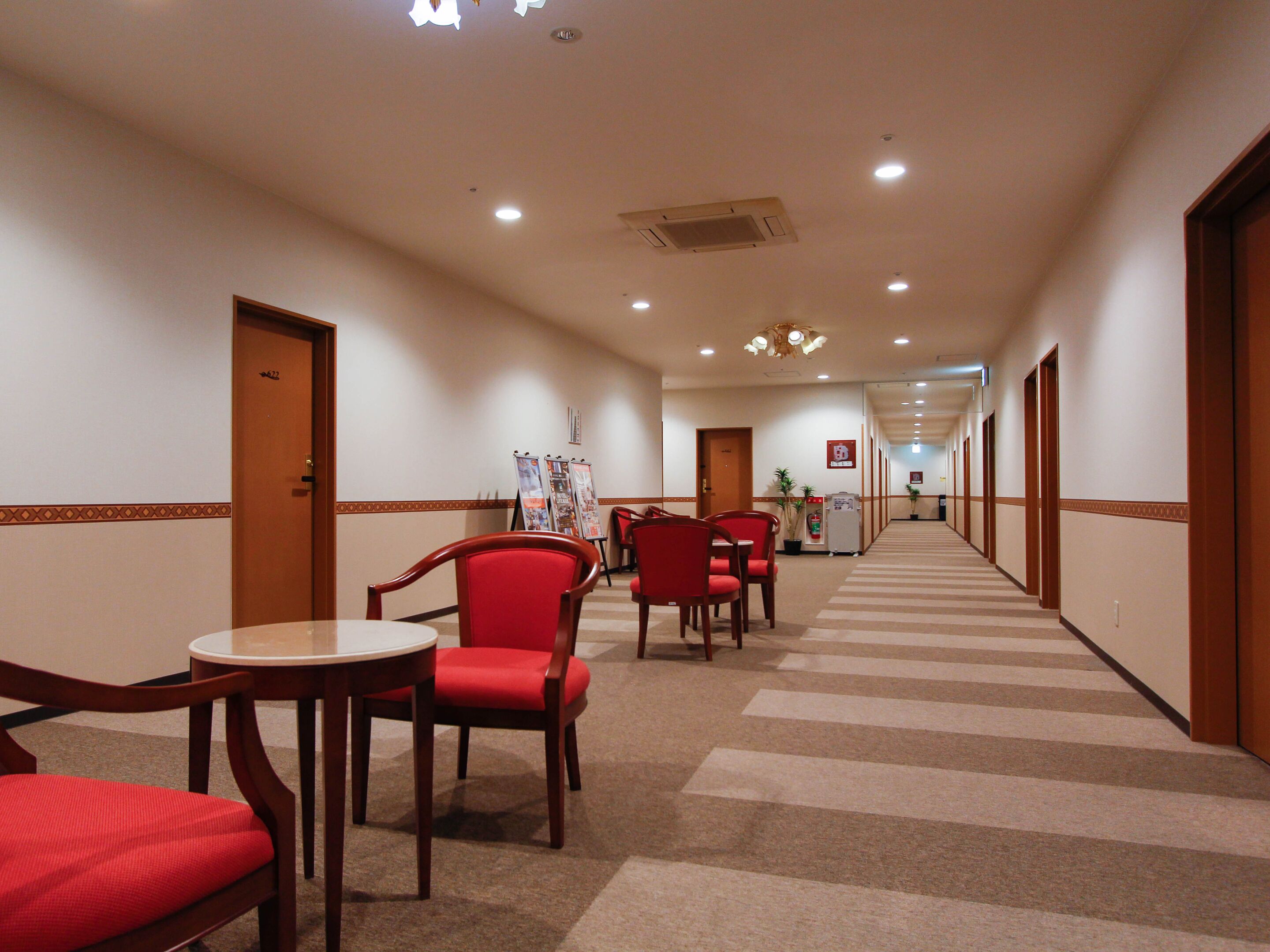 A hotel corridor with a carpeted floor, wooden doors, and red chairs and tables.