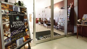 Ein Hotel Eingang mit Glastür, einer Rezeption und einem Plakat, das Hotel LIVEMAX bewirbt.