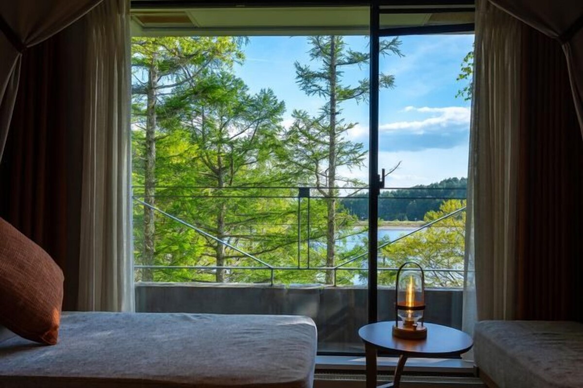 A room with a bed, a small table with a lamp, and a view of trees and a lake through a large window.