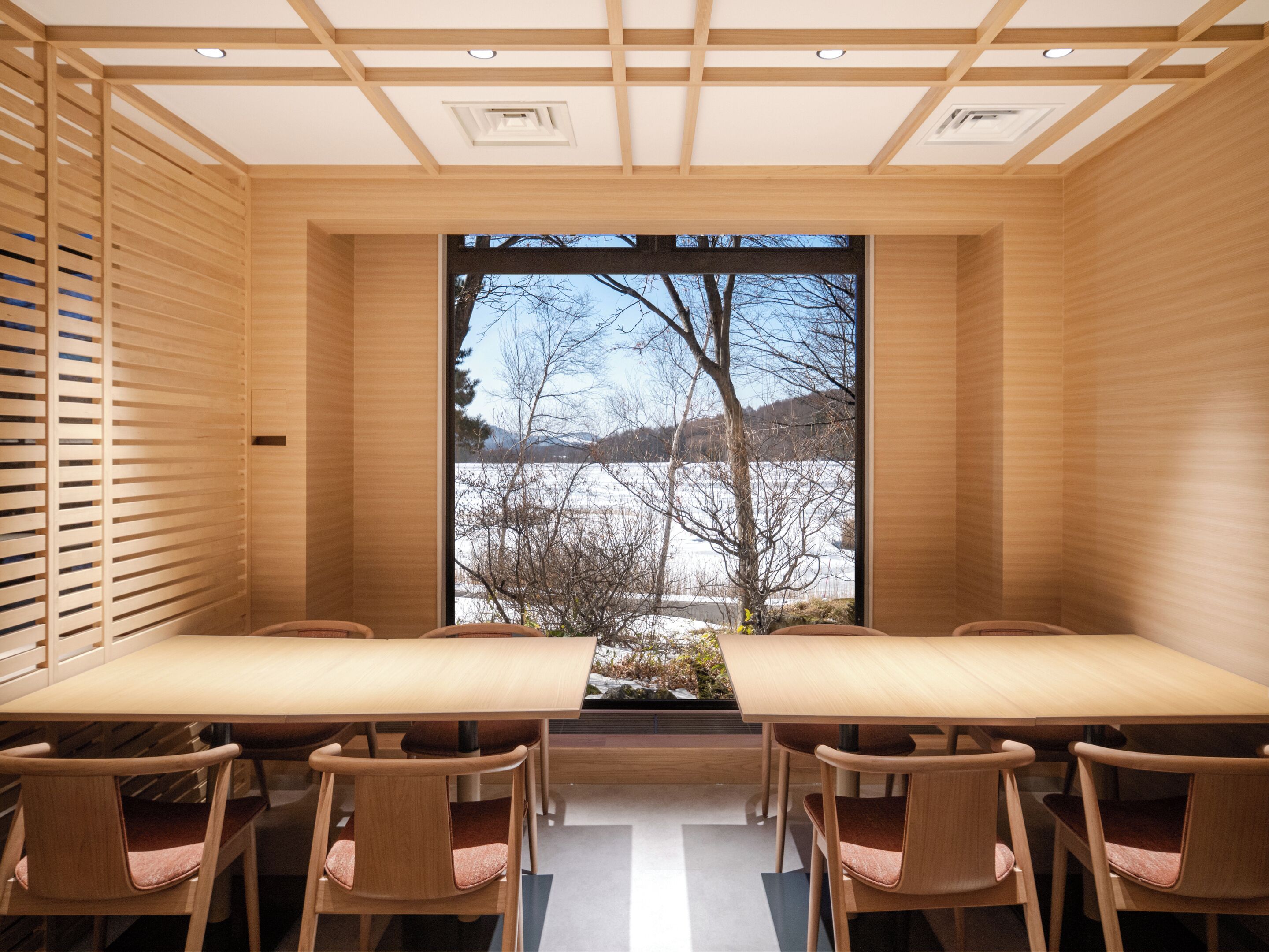A modern interior with a large table and chairs, wooden paneling, and a view of a snowy landscape through a large window.