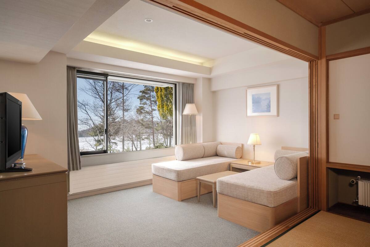 Ein modernes Hotelzimmer mit einer Couch, einem kleinen Tisch, einem Fernseher und einem großen Fenster mit Blick auf Bäume und ein Gebäude.