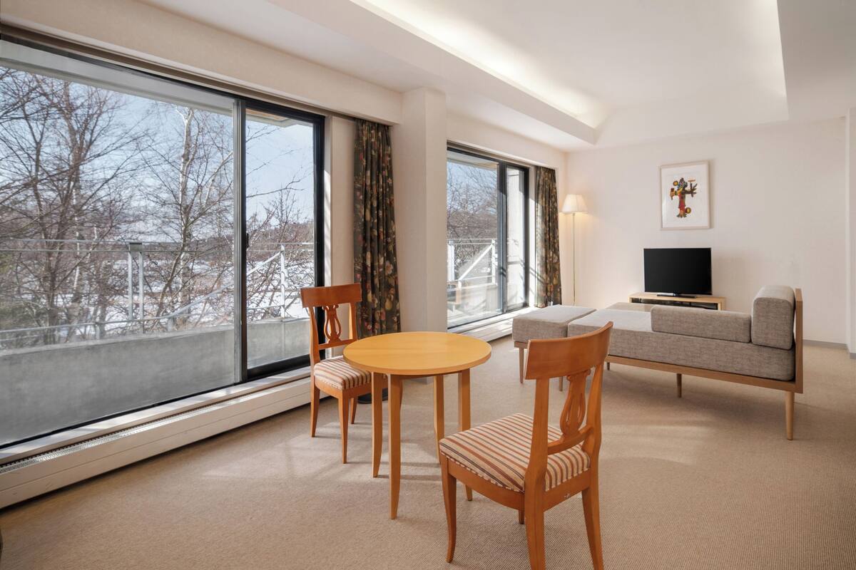 Ein modernes Hotelzimmer mit großem Fenster, einem runden Tisch mit Stühlen, einer Couch und einem Flachbildfernseher.