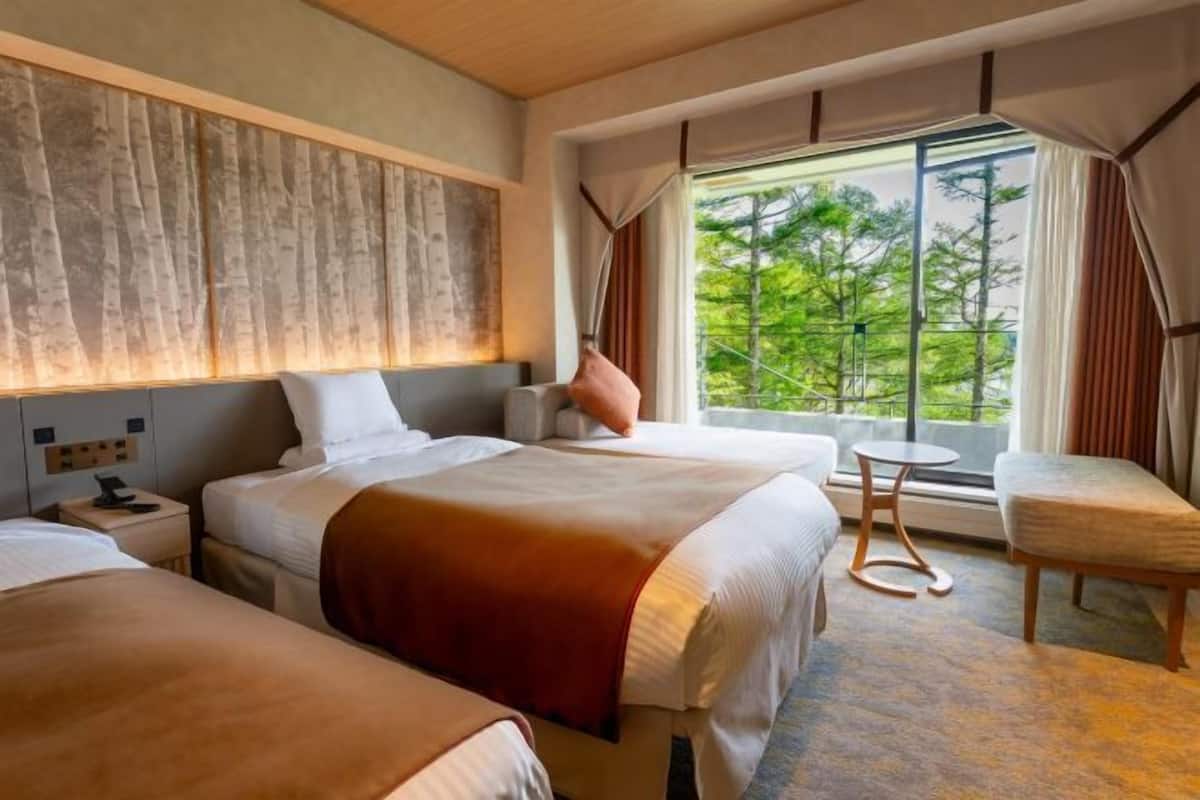 A hotel room with two beds, a small table, a chair, and a large window with a view of greenery.